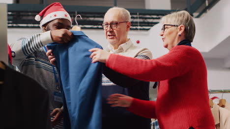 Asistente-Minorista-En-Navidad-Adorna-Una-Boutique-De-Moda-Ayudando-A-Una-Pareja-De-Ancianos-Durante-Su-Frenesí-De-Compras-Navideñas.-Trabajador-Que-Ofrece-Asesoramiento-A-Clientes-De-Edad-Avanzada-Durante-La-Temporada-Festiva