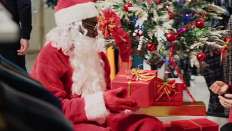 Customer-in-fashion-store-talking-with-actor-dressed-as-Santa-Claus,-sitting-next-to-Christmas-tree-during-festive-promotional-event.-Client-receiving-present-from-worker-during-holiday-season