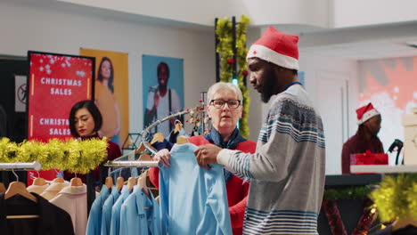 African-american-worker-wearing-Santa-hat-helping-woman-browsing-for-clothes-in-festive-Christmas-decorated-clothing-store.-Employee-assisting-elderly-customer-in-festive-adorn-fashion-shop