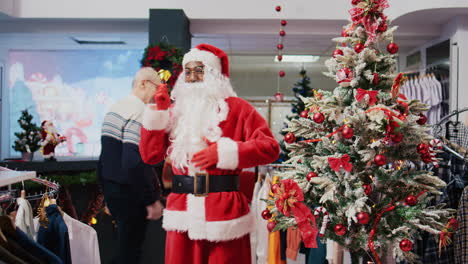 Close-up-shot-of-employee-dressed-as-Santa-Claus-in-Christmas-themed-shopping-mall-clothing-shop.-Retail-assistant-jingling-xmas-bells-and-greeting-clients-in-festive-decorated-fashion-boutique