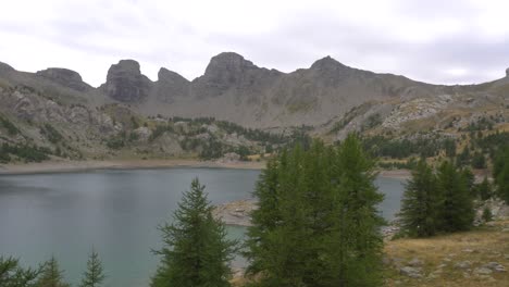 Allos-Lake,-Biggest-Altitude-Lake-of-Europe,-4K-Pan-on-a-Cloudy-Day,-Showing-Blue-Water-Surrounded-by-Grey-Peaks-and-Pine-Trees