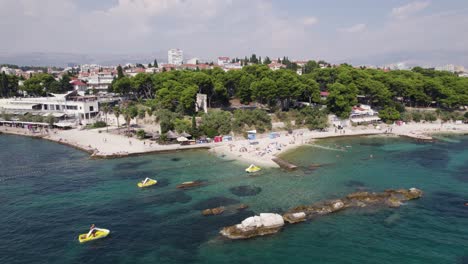 Aerial:-Ovcice-Beach,-Split,-with-boats,-rocks,-and-distant-town-backdrop