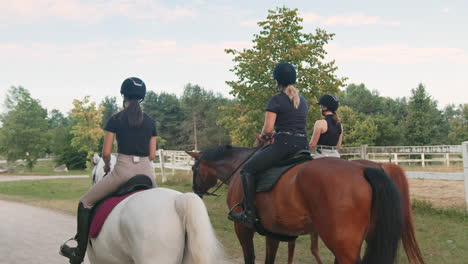 Three-female-riders-riding-horses-side-by-side-near-wood-fencing,-rear-view