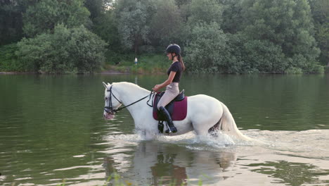 Girl-riding-a-snow-white-horse-down-the-calm-river-water,-tracking-shot
