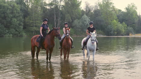 Three-women-enjoying-equestrian-leisure-and-river,-sitting-on-horses,-wide-shot