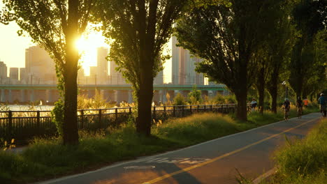 Sun-Setting-Behind-Buildings-View-from-Seoul-Hangang-or-Han-River-Park,-People-Cycling-on-Bicycles,-Back-of-Delivery-Man-Riding-Electric-Bike-on-Riverside-Track