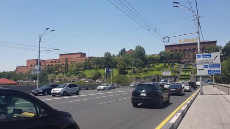 Traffic-in-Downtown-Yerevan,-Armenia,-Cars-on-Victory-Bridge-With-Ararat-Brandy-Factory-in-Background