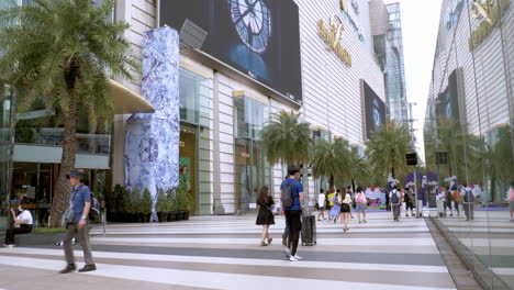 People-are-busy-walking-outside-of-Siam-Paragon-in-Bangkok-with-the-BTS-train-station-with-trains-leaving-and-stopping-for-passengers,-Thailand