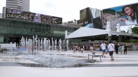 Ein-Wasserbrunnen-Vor-Siam-Paragon-Und-Der-Siam-BTS-Station-Mit-Einem-Wartenden-Zug-Und-Menschen,-Die-Herumlaufen,-Bangkok,-Thailand