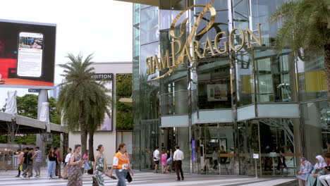 La-Gente-Está-Ocupada-Caminando-Fuera-De-Siam-Paragon-En-Bangkok-Con-La-Estación-De-Tren-BTS-Con-Trenes-Que-Salen-Y-Paran-Para-Pasajeros,-Tailandia