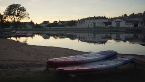 Paddle-boards-sit-idle-next-to-a-peaceful-river-and-small-village-as-evening-closes-in