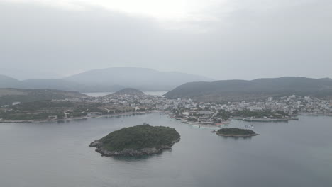 Drone-shot-flying-over-the-water-to-the-small-islands-in-front-of-Ksamil-Albania-in-the-morning-on-a-cloudy-grey-day-with-boats-around-and-haze-in-the-sky-LOG