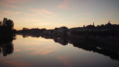 Golden-hour-skyline-above-tranquil-river-water-next-to-old-quaint-village