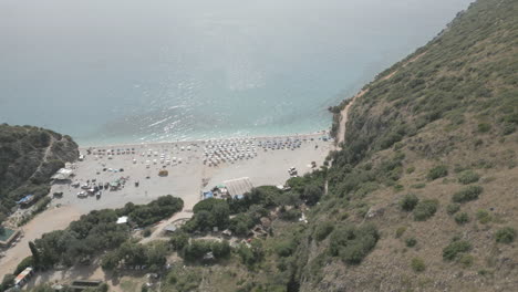 Drone-shot-over-Gjipe-beach-Albania-with-tourists-on-the-beach-and-clear-blue-water-near-the-mountains-and-the-sea-on-a-sunny-and-bright-day-and-green-nature-around-with-sun-beds-towels-LOG