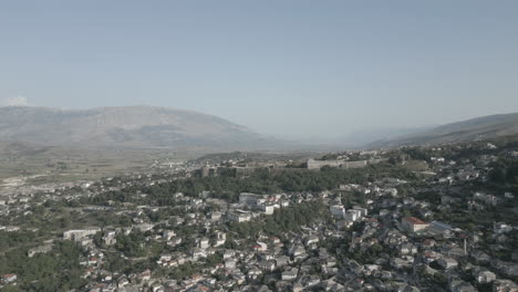 Drone-shot-above-the-city-Gjirokaster-Albania-with-the-castle-on-the-mountain-on-a-sunny-day-with-haze-in-the-air-and-the-city-of-white-houses-underneath-and-mountains-in-the-background-LOG