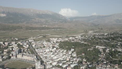 Drone-shot-above-the-city-Gjirokaster-Albania-on-a-sunny-day-with-haze-in-the-air-and-the-city-of-white-houses-underneath-and-mountains-and-clouds-in-the-background-LOG