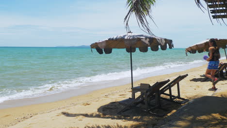 Grandes-Sombrillas-Y-Sillas-De-Playa-En-Un-Resort-En-Pattaya-Mientras-Una-Mujer-Se-Acerca-Al-Agua-Para-Nadar-Mientras-Las-Olas-Llegan-A-La-Orilla.