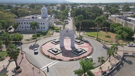 Toma-Aérea-Hacia-Atrás-Del-Arco-Triunfal-Y-Vehículo-De-Conducción-En-La-Rotonda-Durante-El-Día-Soleado---San-Juan,-República-Dominicana