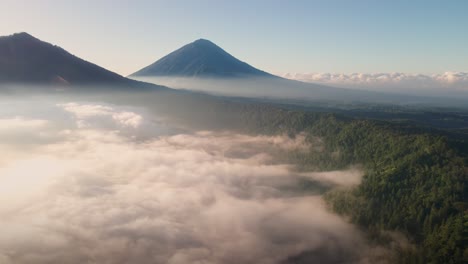 Aerial-view-rising-over-a-misty-valley-toward-Volcanic-peaks-of-sunny-Bali,-Asia