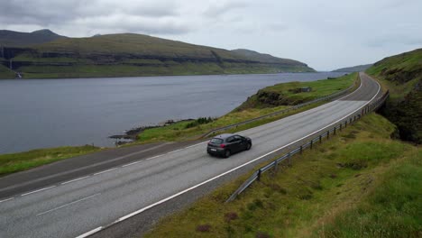 Aerial-drone-starts-tracking-a-car-driving-through-an-Eysturoy-road,-Faroe-Islands