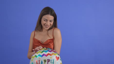 A-young-woman-poses-with-a-Happy-Birthday-balloon,-isolated-on-a-blue-studio-background