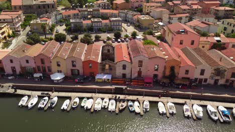 Bosa-colorful-houses-facing-the-river-Temo-with-boats-docked,-sideways,-aerial