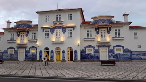 Woman-walks-in-front-of-illuminated-Aveiro-old-train-station,-tracking-shot