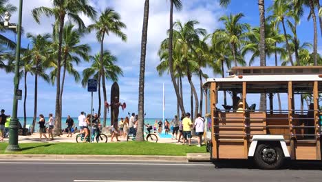 Streetcar-passes-Waikiki,-Hawaii-beach-with-tourists-and-palm-trees-on-sunny-day