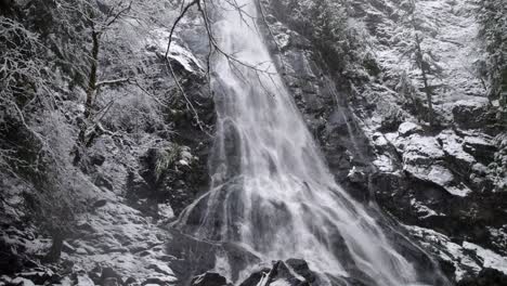 Snow-gently-falls-while-orbiting-slowly-through-a-winter-forest,-cascading-waterfall,-aerial-drone