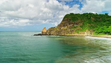 Sunny-beach-and-rocky-cliffs-at-San-Juan-del-Sur,-Nicaragua,-aerial-forward