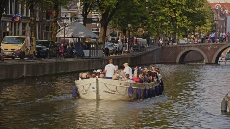Plano-Amplio-De-Un-Hermoso-Canal-En-El-Centro-De-La-Ciudad-De-Amsterdam-Con-Un-Barco-Turístico-Lleno-De-Turistas-Navegando-Sobre-El-Agua