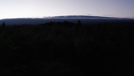 Low-aerial-shot-flying-over-treetops-towards-towering-Mauna-Loa-peeking-above-the-clouds-at-sunset-on-the-island-of-Hawai'i