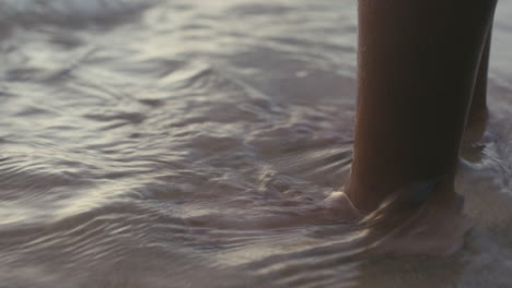 Close-up-of-water-washing-over-child's-feet-standing-on-sand-on-the-beach
