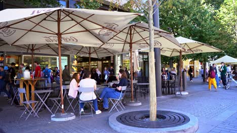 Panorama-De-Gente-Sentada-Hablando-En-Un-Restaurante-Con-Sombrillas-Gigantes-En-Medio-De-Las-Calles-Con-Turistas-Que-Pasan-Constantemente-En-Un-Día-Soleado-En-Praga,-República-Checa