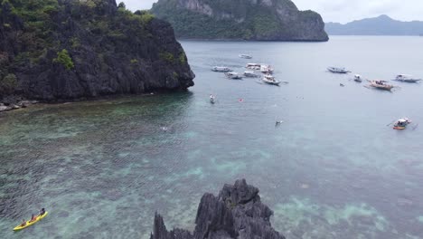 Flying-over-Tourists-kayaking-and-swimming-from-anchored-island-hopping-tour-Boats-to-Cadlao-Lagoon-or-Ubugon-Cove,-El-Nido,-Aerial-dolly-in
