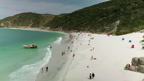 Aerial-flyover-sandy-beach-with-resting-tourist-and-clear-green-ocean-water-in-summer---Arraial-do-Cabo,-Brazil