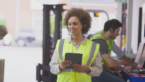 Female-Manager-Wearing-Headset-In-Logistics-Distribution-Warehouse-Using-Digital-Tablet