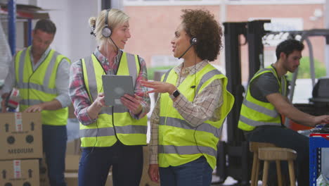 Two-Female-Workers-Wearing-Headsets-In-Logistics-Distribution-Warehouse-Using-Digital-Tablet