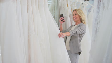 Mature-Woman-In-Bridal-Wedding-Dress-Shop-Taking-Photo-On-Mobile-Phone