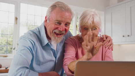Retired-Senior-Couple-In-Kitchen-At-Home-Making-Video-Call-Using-Laptop