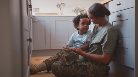 Amerikanische-Armeemutter-In-Uniform-Zu-Hause-Auf-Urlaub-Umarmt-Sohn,-Der-Auf-Dem-Boden-In-Der-Familienküche-Sitzt