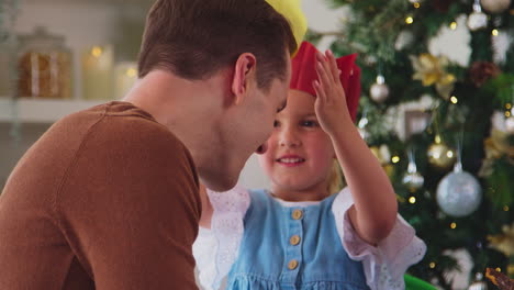 Hija-Poniéndole-Un-Sombrero-De-Papel-Al-Padre-Como-Familia-Celebrando-La-Navidad-En-Casa-Con-Comida