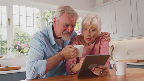Rentnerpaar-Sitzt-Zu-Hause-In-Der-Küche,-Trinkt-Kaffee-Und-Nutzt-Ein-Digitales-Tablet