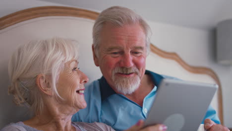 Lächelndes-Rentnerpaar-Liegt-Zu-Hause-Im-Bett-Und-Schaut-Gemeinsam-Auf-Ein-Digitales-Tablet