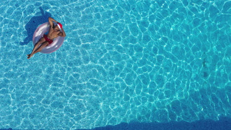 Drone-Shot-Of-Man-On-Christmas-Holiday-Floating-On-Inflatable-Ring-In-Swimming-Pool-In-Santa-Hat