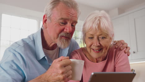 Primer-Plano-De-Una-Pareja-De-Ancianos-Jubilados-Sentados-En-La-Cocina-De-Casa-Tomando-Café-Y-Usando-Una-Tableta-Digital