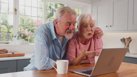 Retired-Senior-Couple-In-Kitchen-At-Home-Making-Video-Call-Using-Laptop