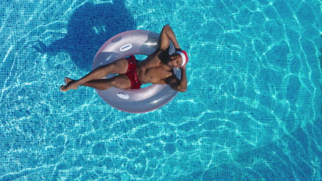 Drone-Shot-Of-Man-On-Christmas-Holiday-Floating-On-Inflatable-Ring-In-Swimming-Pool-In-Santa-Hat