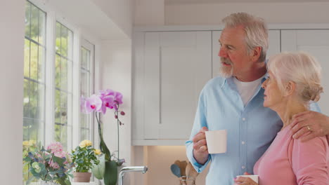 Liebevolles-älteres-Ehepaar-Im-Ruhestand,-Das-Zu-Hause-Am-Fenster-In-Der-Küche-Steht-Und-Kaffee-Trinkt