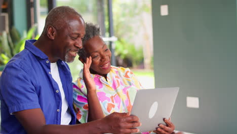 Smiling-Senior-Couple-Sitting-At-Home-Making-Video-Call-On-Laptop-Computer